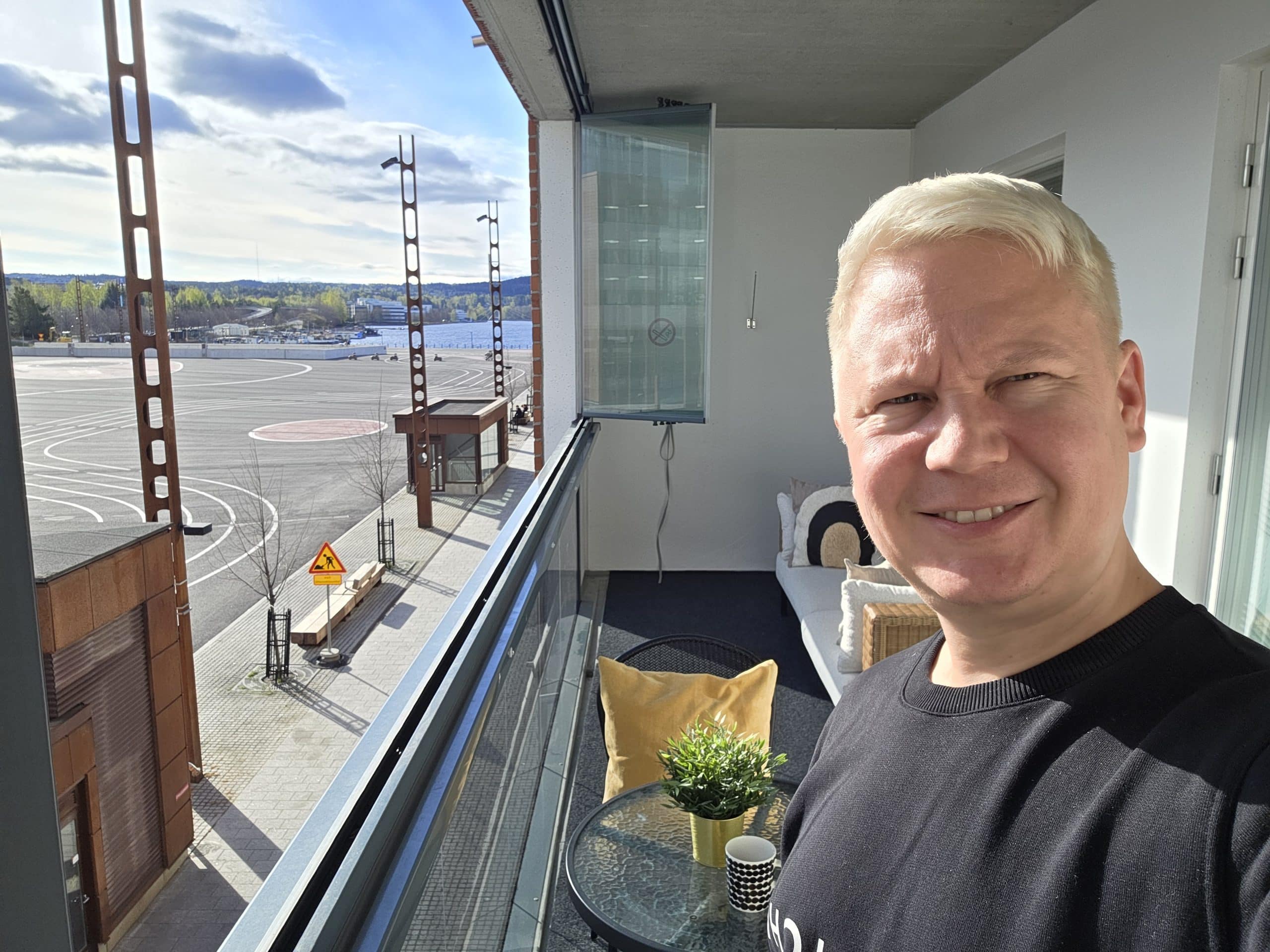 Selfie of a man on a balcony overlooking a construction site and water, with blue sky and distant hills in the background.